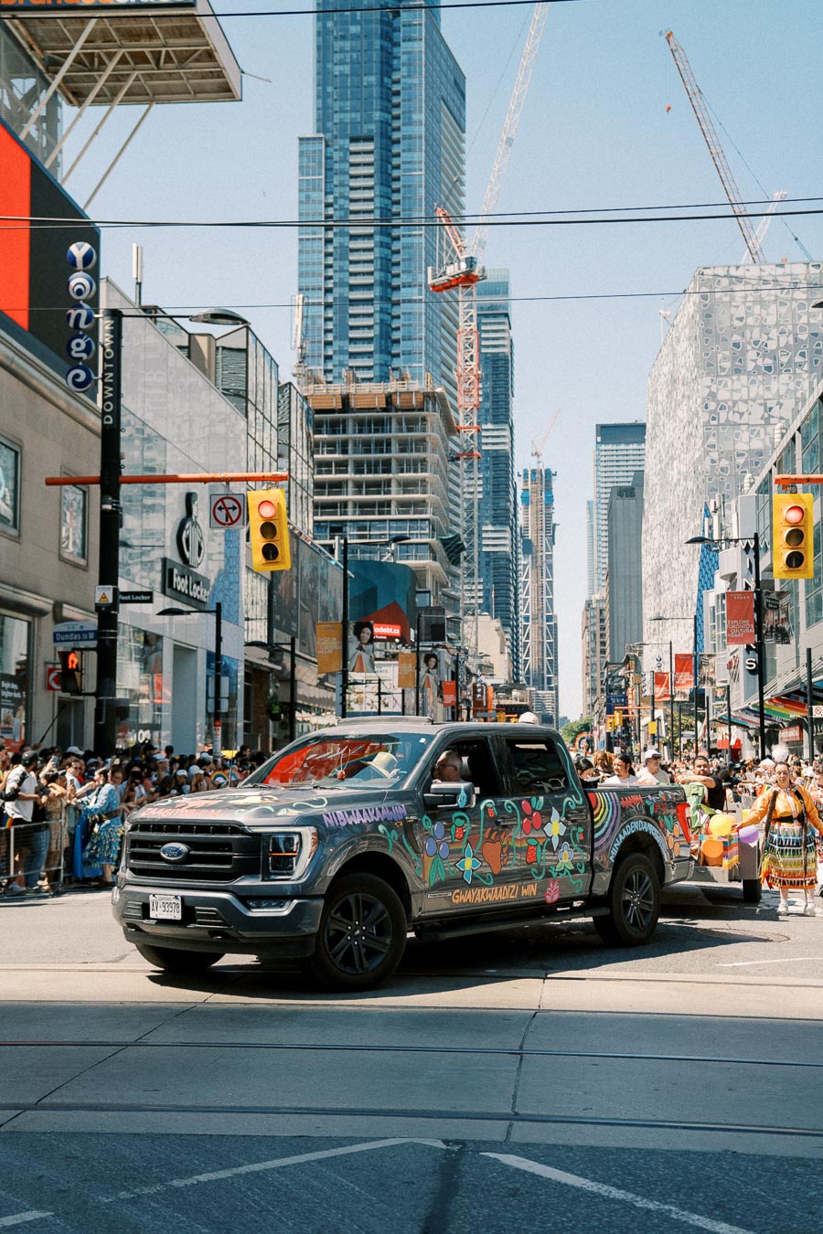 A decorated Ford truck participates in a city parade, set against a backdrop of skyscrapers and construction cranes, indicating urban development.
