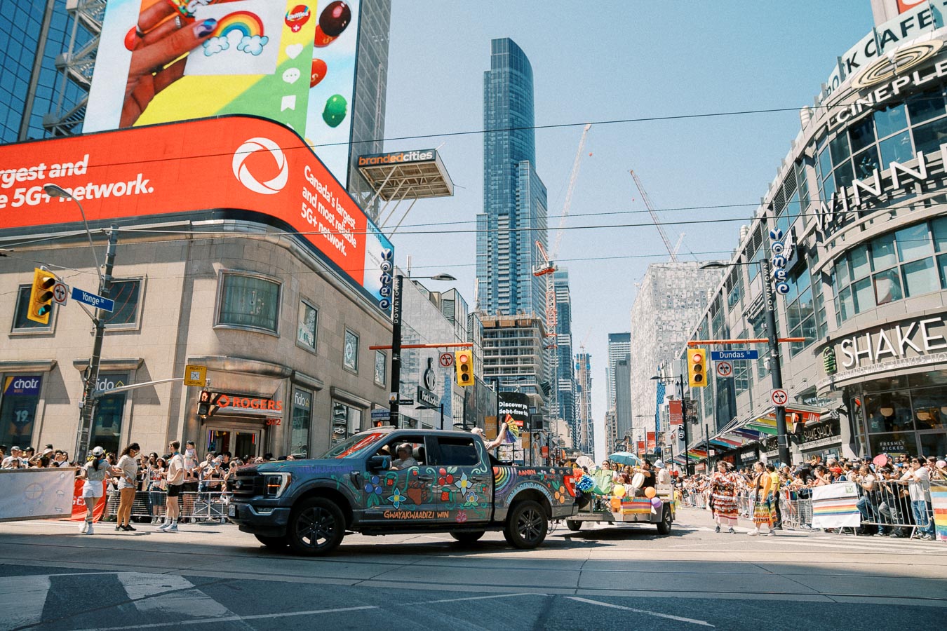 A vibrant parade in downtown Toronto, featuring a decorated truck and participants in traditional attire, passes through the bustling intersection of Yonge and Dundas Streets.