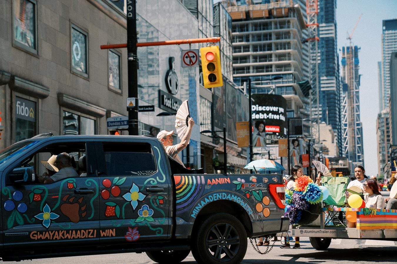 A vibrantly decorated pickup truck participates in a city parade, with passengers holding fans and umbrellas. The street is lined with tall buildings and a traffic light, indicating an urban setting.
