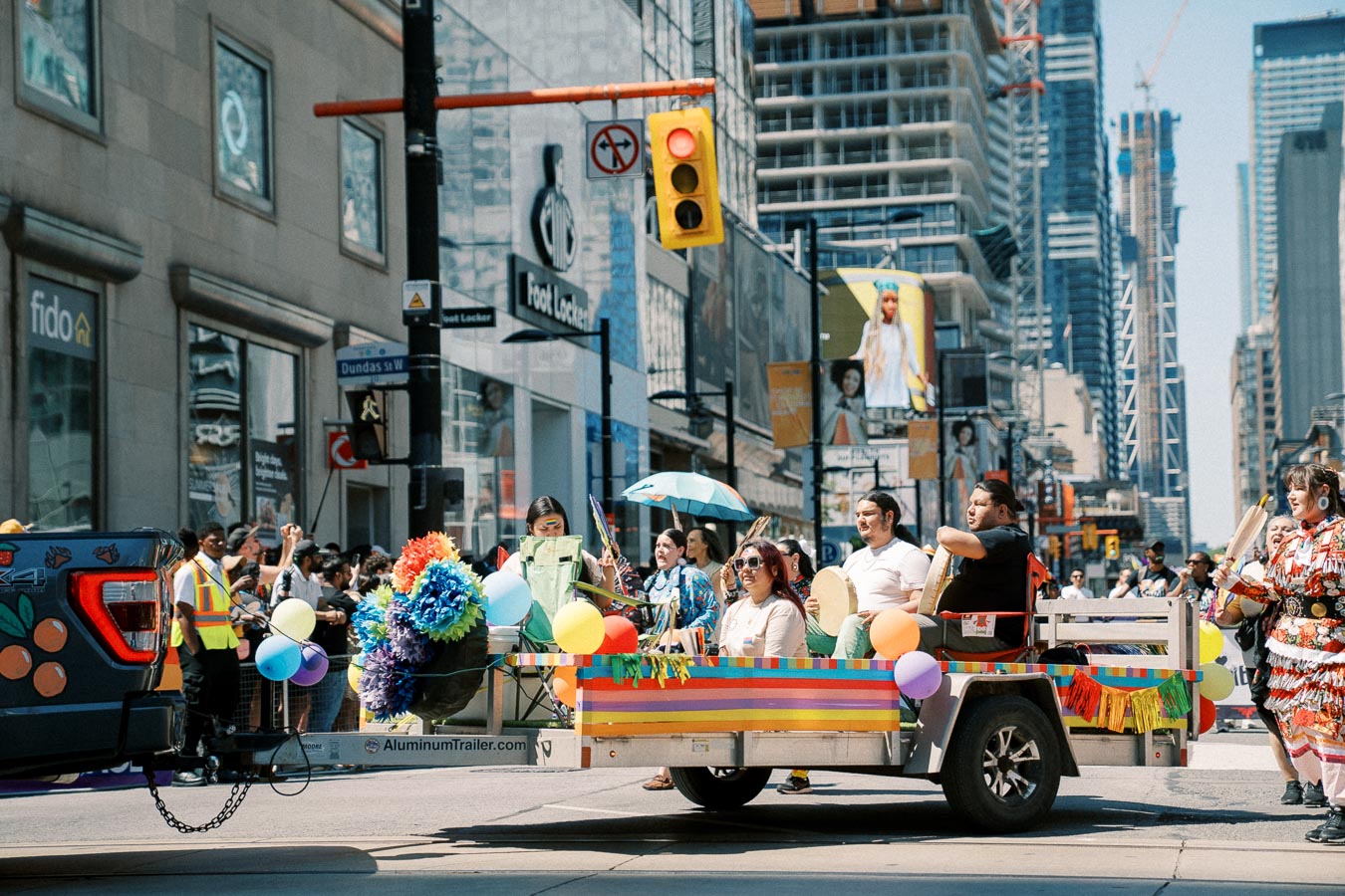A vibrant parade float adorned with balloons and flowers makes its way through a bustling city street, with participants holding drums and umbrellas. The scene is set against a backdrop of tall buildings and a crowd of onlookers.