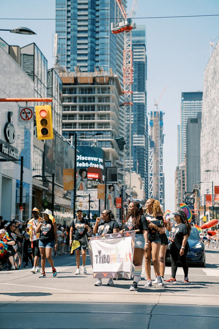 A vibrant parade takes place in a bustling city street, with participants holding a banner and surrounded by tall buildings and construction cranes.