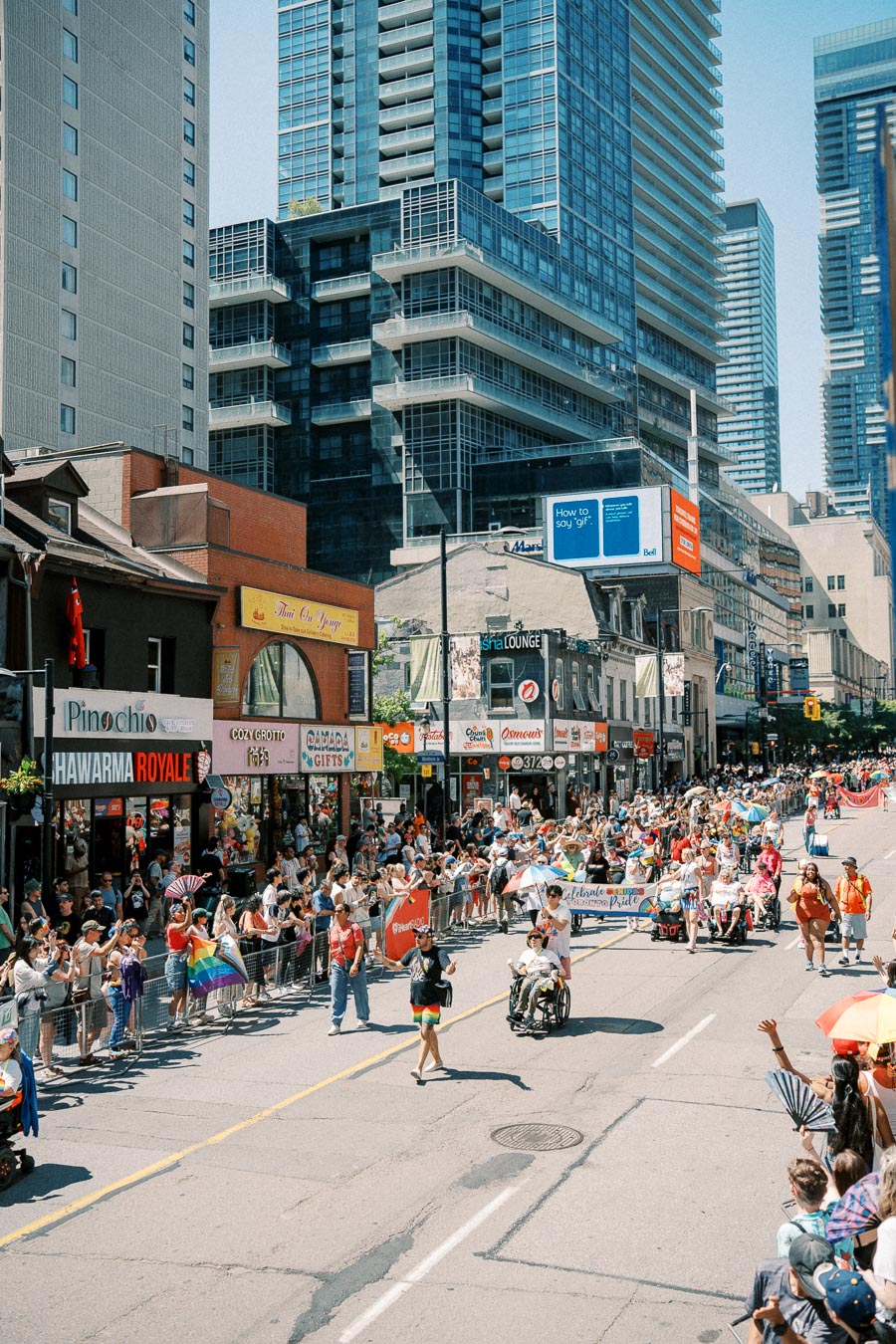 A lively street parade with participants and spectators, featuring colorful outfits and flags, set against a backdrop of urban buildings and storefronts.