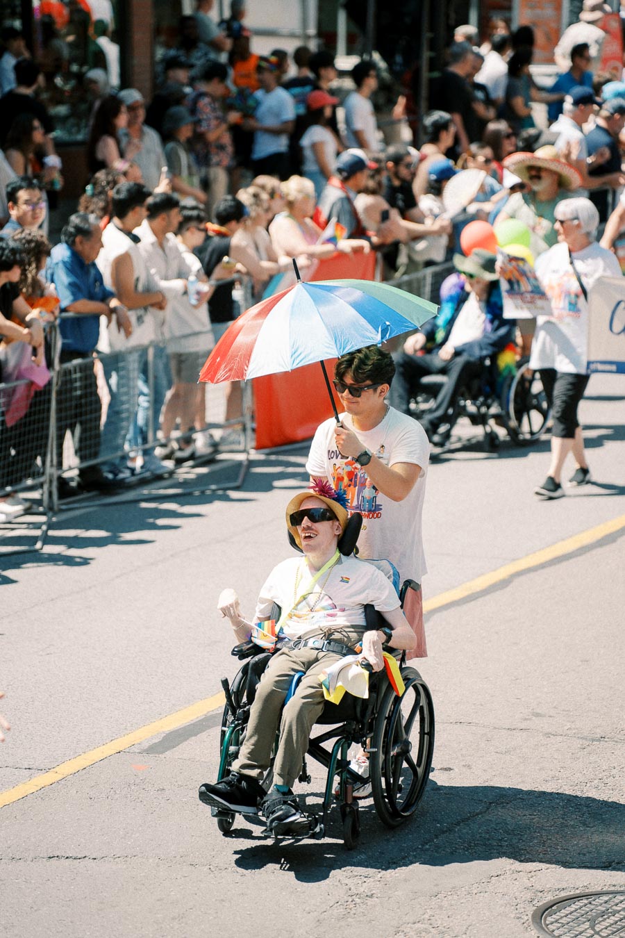 A parade participant in a wheelchair is pushed by another person holding a colorful umbrella, surrounded by a crowd.