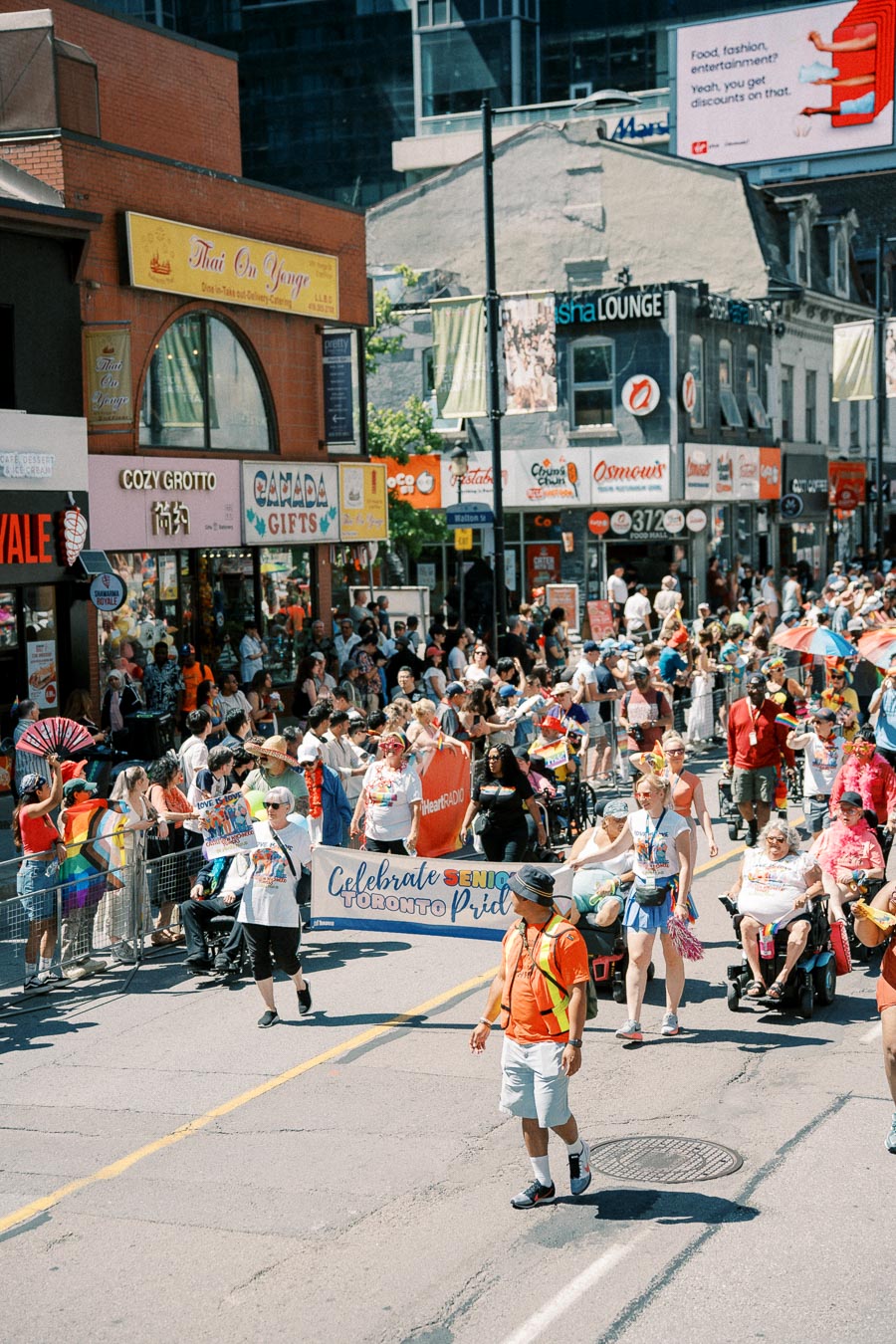 Participants in a Toronto Pride parade celebrate with a banner reading 'Celebrate Seniors Toronto Pride', surrounded by spectators and local shops.