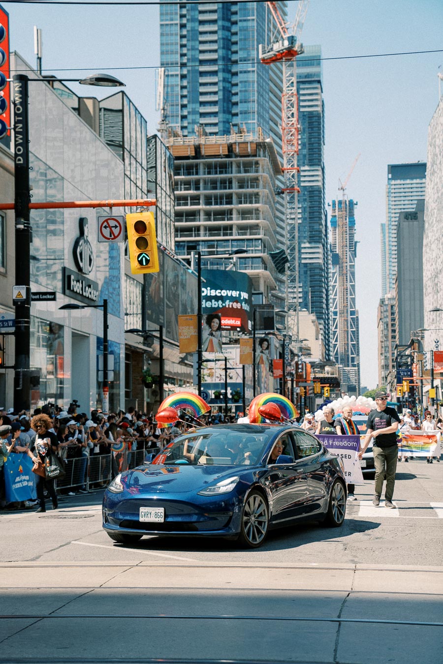 A blue Tesla adorned with rainbow balloons drives through a city parade, surrounded by skyscrapers and a cheering crowd.