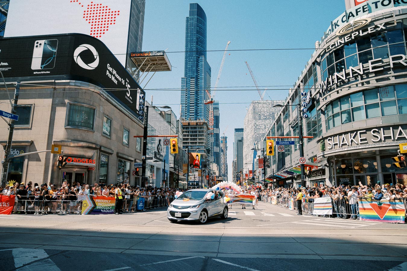 A vibrant parade takes place on a bustling street in Toronto, featuring a silver car adorned with balloons and a large crowd of spectators. The scene is set against a backdrop of towering skyscrapers and prominent billboards.