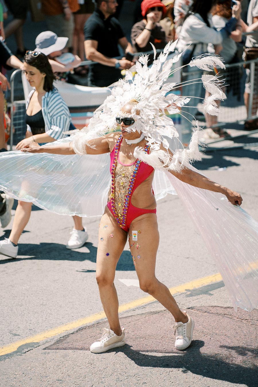 A person dressed in a vibrant costume adorned with feathers and sequins participates in a parade, surrounded by onlookers.