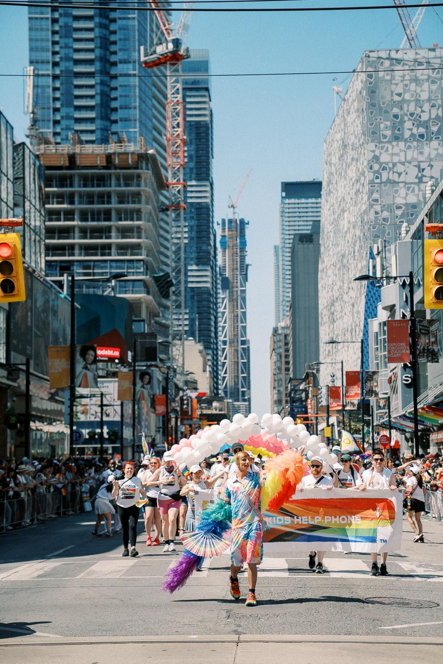 Participants in a pride parade march down a city street, holding a colorful banner and balloons, surrounded by tall buildings and construction cranes.