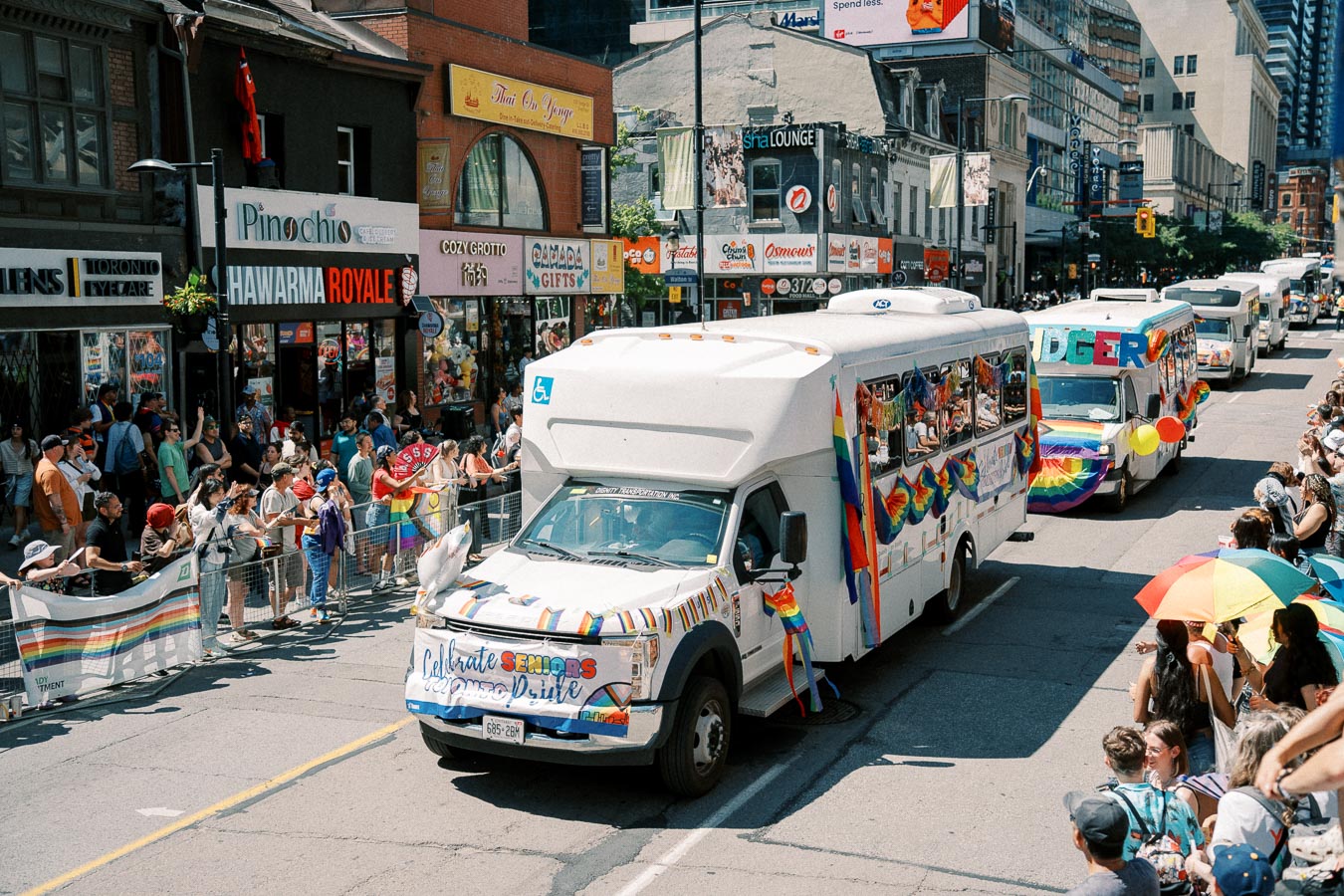A vibrant parade featuring decorated buses and a crowd celebrating on a city street, with rainbow flags and banners.