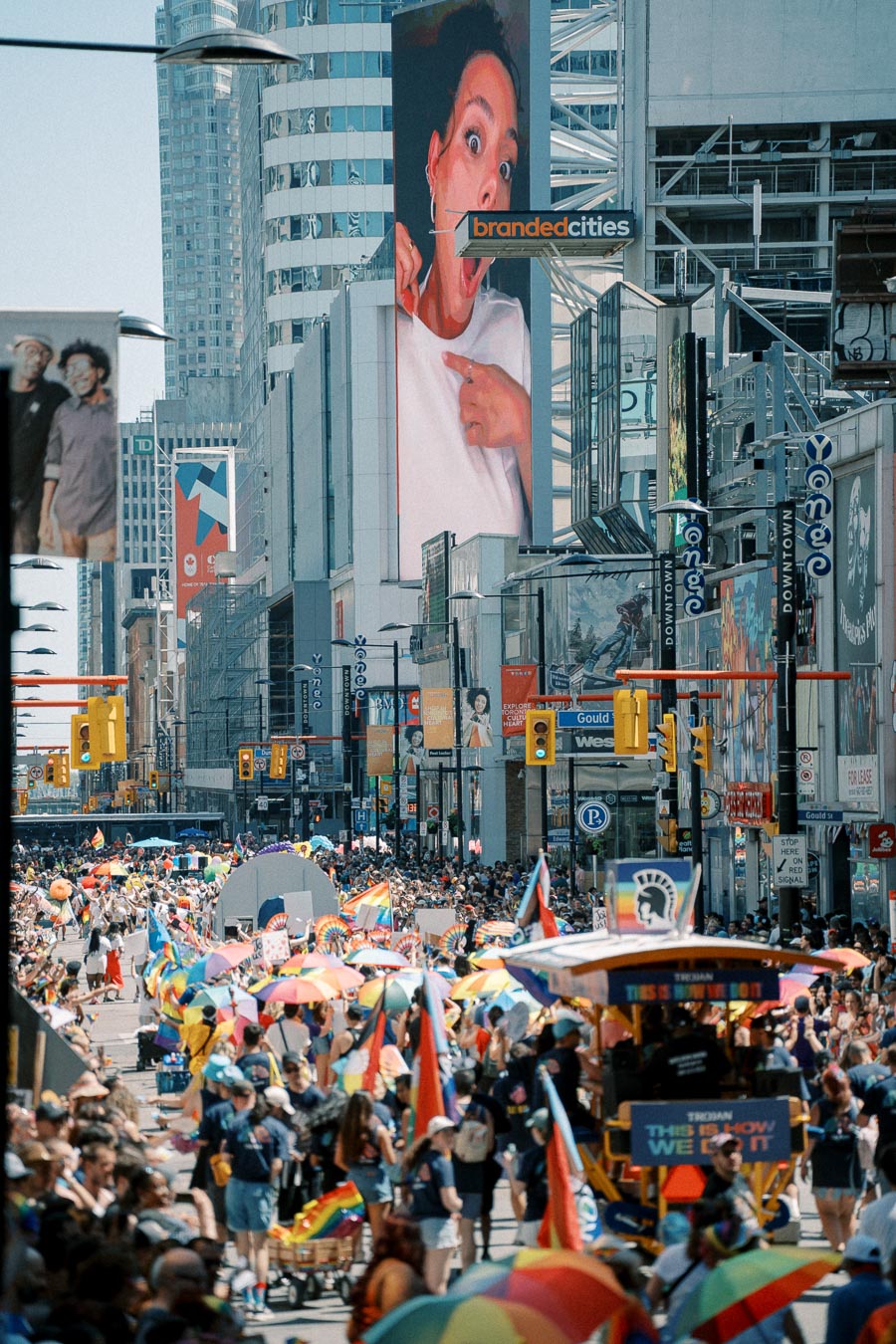 A lively parade takes place on a bustling city street, featuring participants with rainbow flags and umbrellas, set against a backdrop of skyscrapers and digital billboards.
