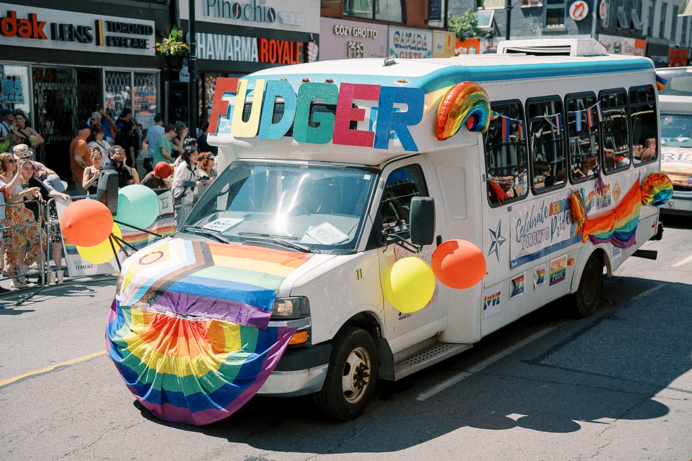 A bus decorated with rainbow flags and balloons participates in a pride parade, featuring the word 'FUDGER' on top.