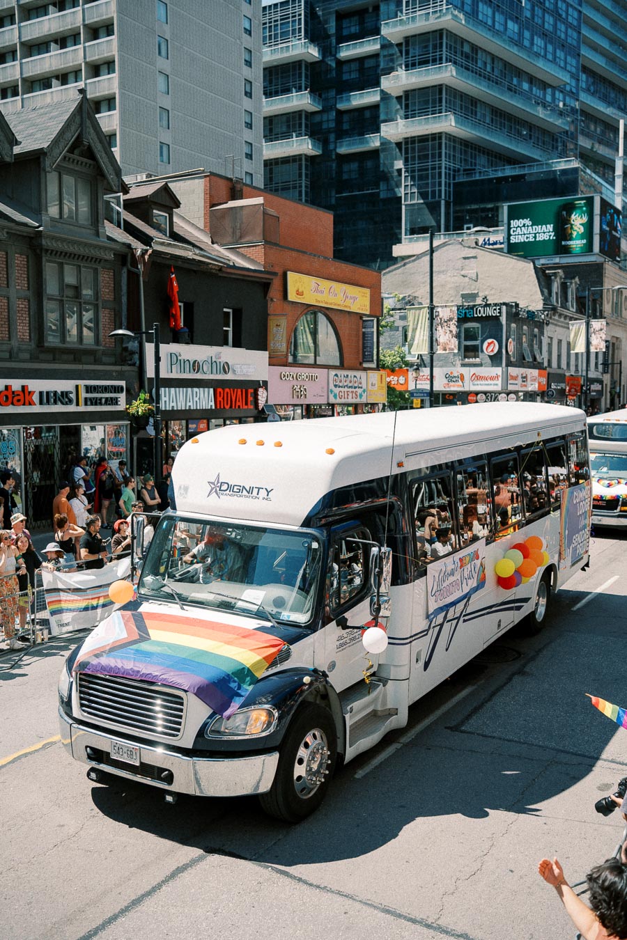 A bus adorned with a rainbow flag and balloons participates in a parade on a city street, surrounded by spectators and shops.