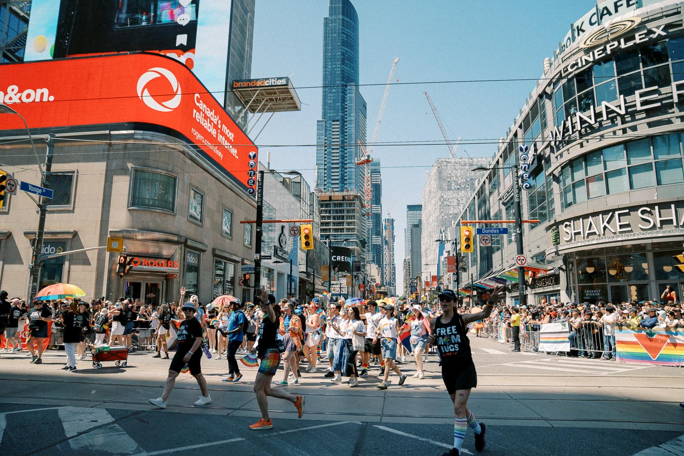 A lively Pride parade at the intersection of Yonge and Dundas in Toronto, featuring colorful participants and spectators, with prominent storefronts and digital billboards in the background.
