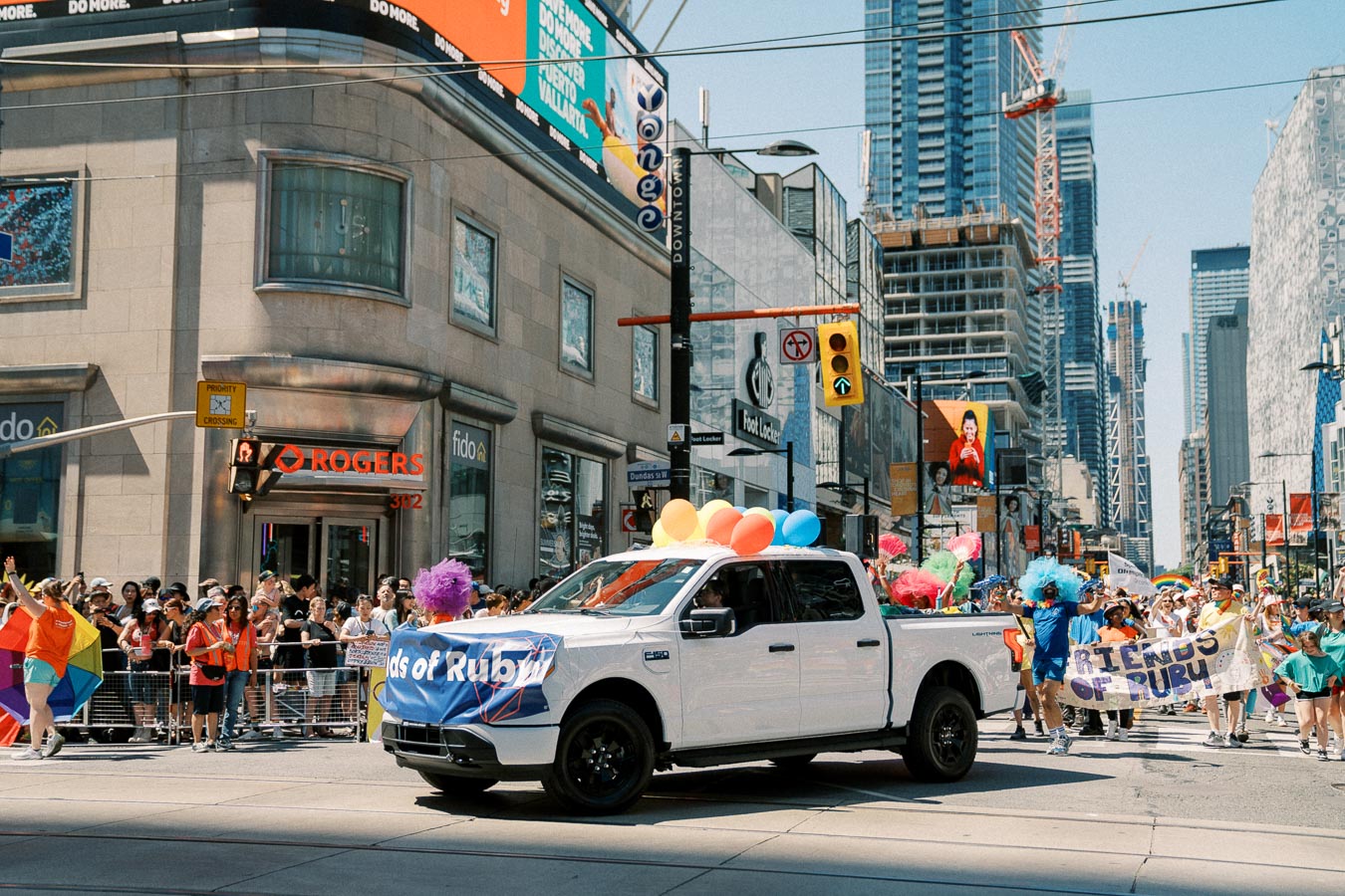 A vibrant pride parade takes place in a bustling city, featuring a white pickup truck adorned with colorful balloons and a banner. Spectators line the street, and tall buildings tower in the background.