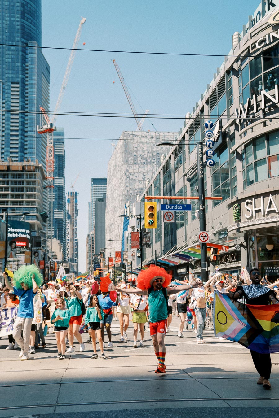 A lively parade on Dundas Street featuring participants in colorful costumes and flags, with urban buildings and construction cranes in the background.