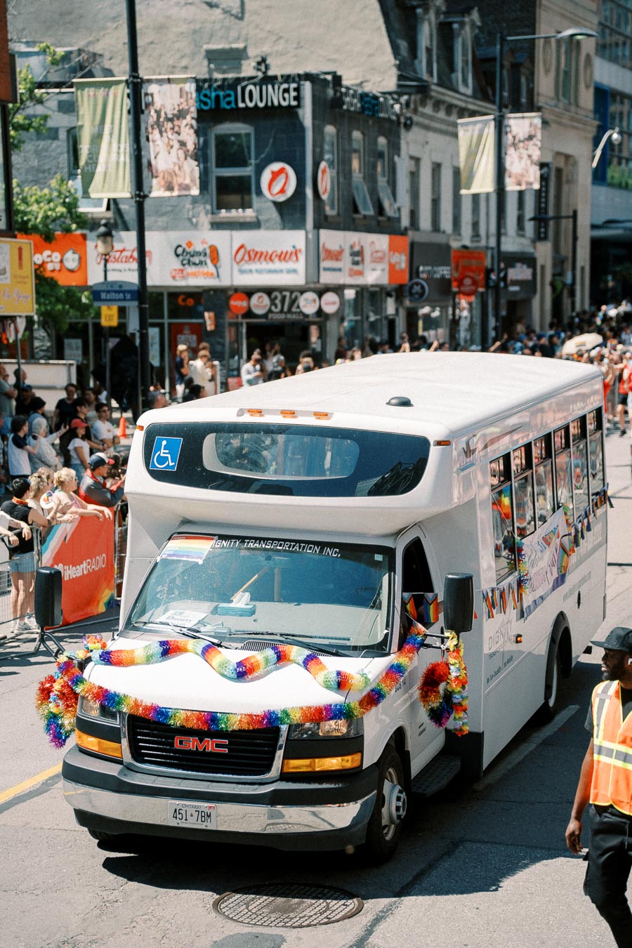 A white GMC bus adorned with rainbow decorations participates in a city parade, surrounded by spectators and vibrant storefronts.