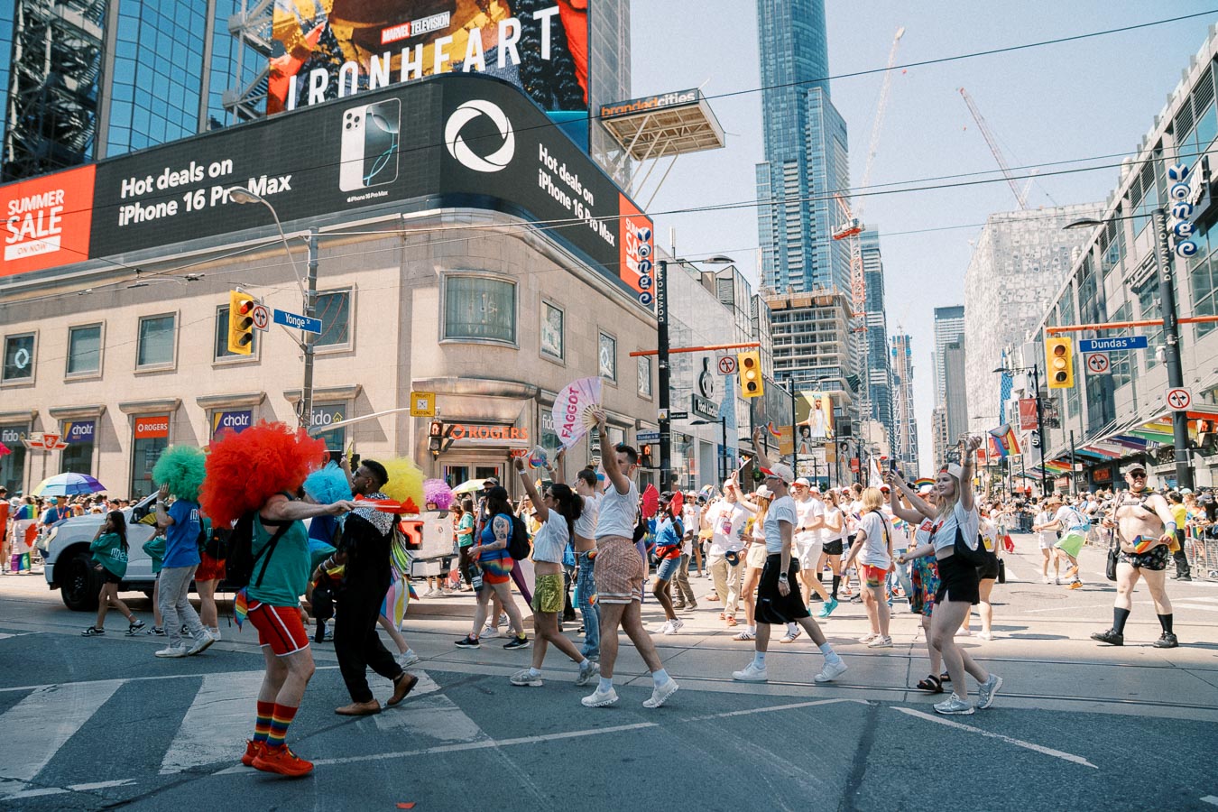 A vibrant parade takes place at the intersection of Yonge and Dundas in Toronto, featuring participants in colorful costumes and wigs, with large digital billboards and skyscrapers in the background.