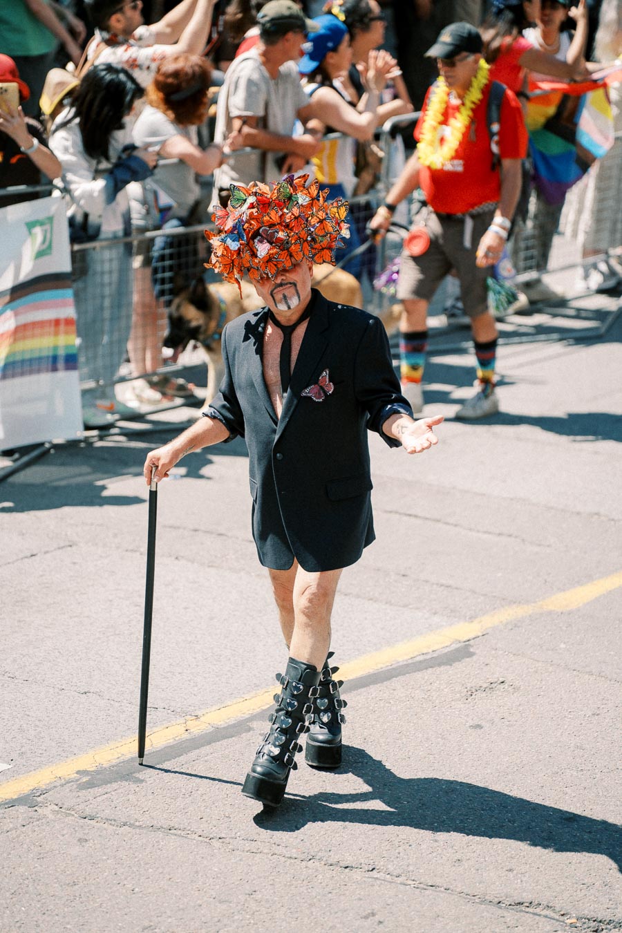 A parade participant wearing a butterfly-themed hat and platform boots walks confidently, holding a cane, during a vibrant street parade.