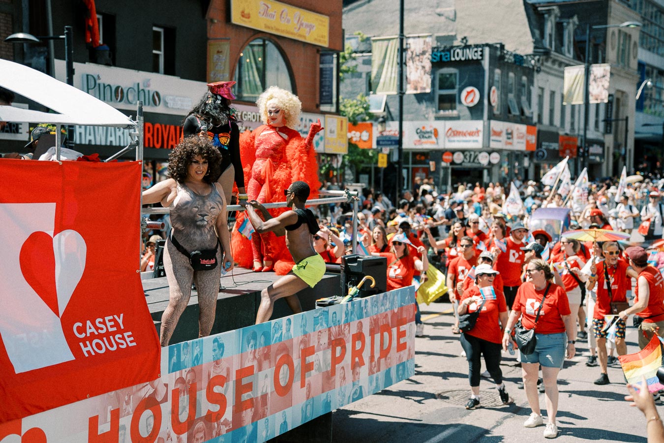 A lively pride parade with a float from 'Casey House' featuring performers in colorful costumes and a crowd of participants in red shirts.