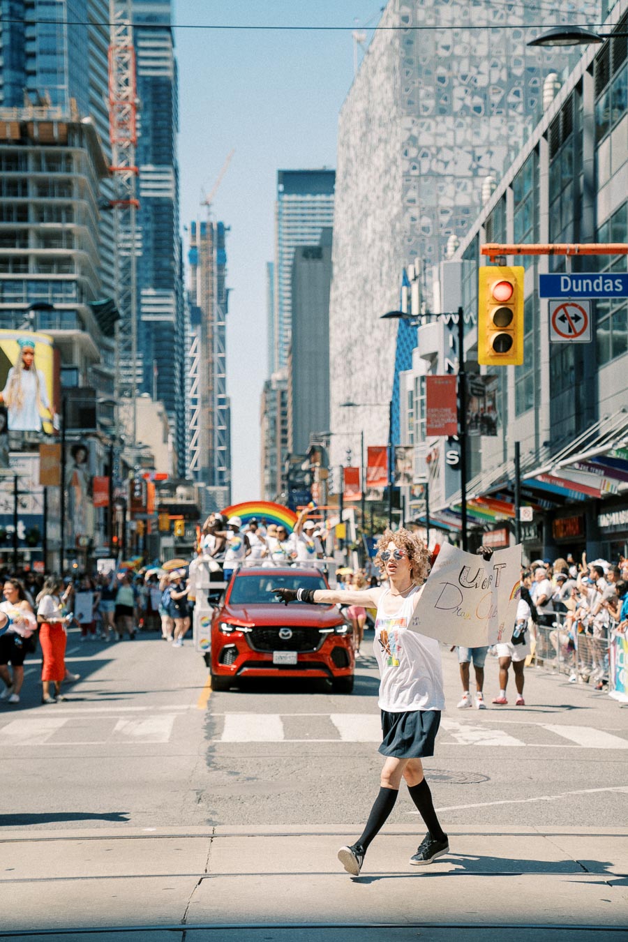 A vibrant parade scene in a city, featuring a person holding a sign and a rainbow float in the background, with tall buildings lining the street.