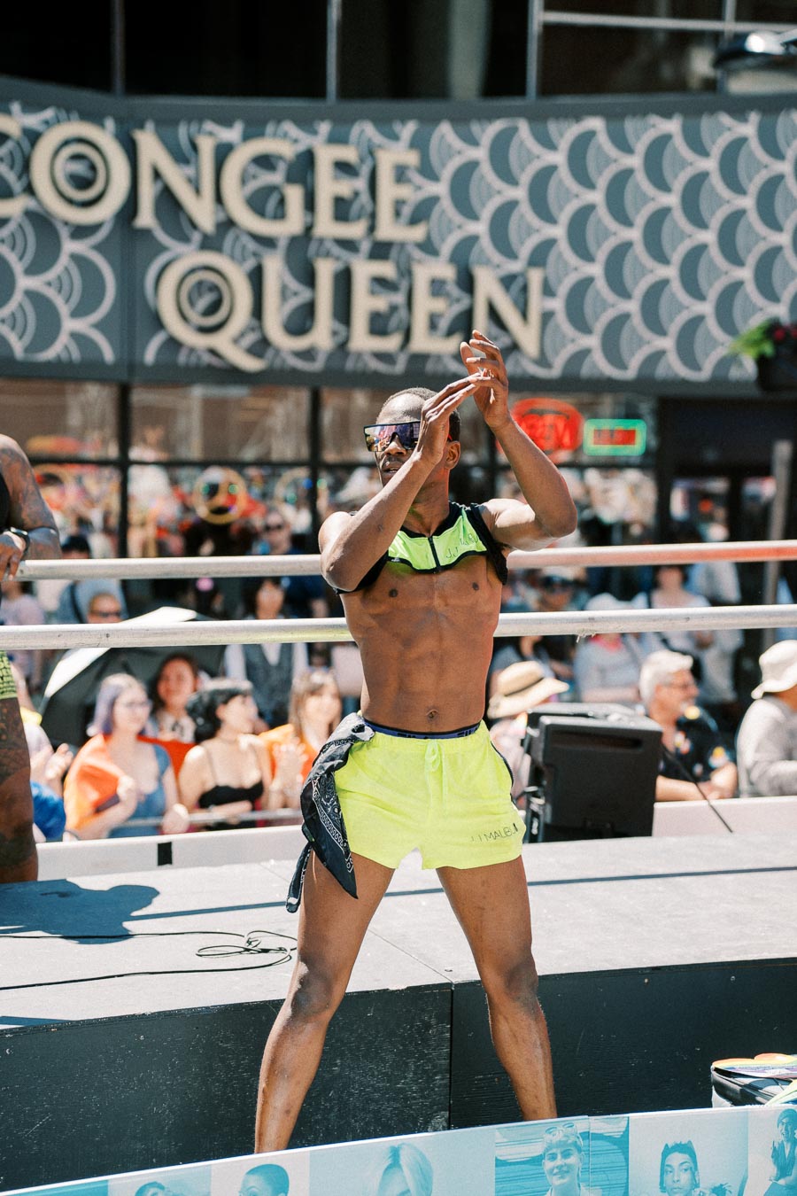 A performer in neon green shorts claps on stage during a lively outdoor event, with an engaged audience in the background.