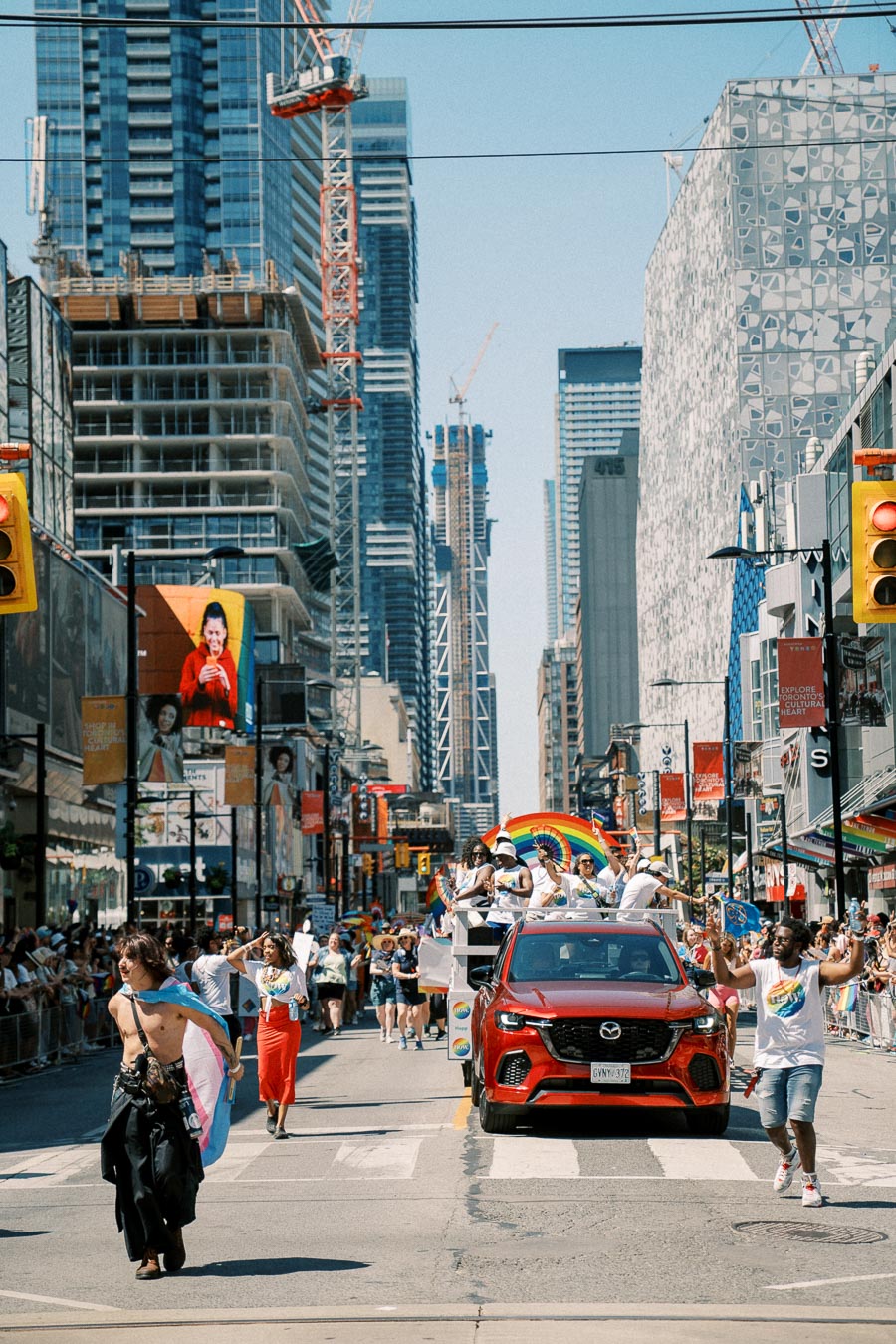 A lively pride parade takes place on a city street, featuring a red Mazda car and participants celebrating with rainbow flags. Skyscrapers and construction cranes are visible in the background, adding to the urban atmosphere.