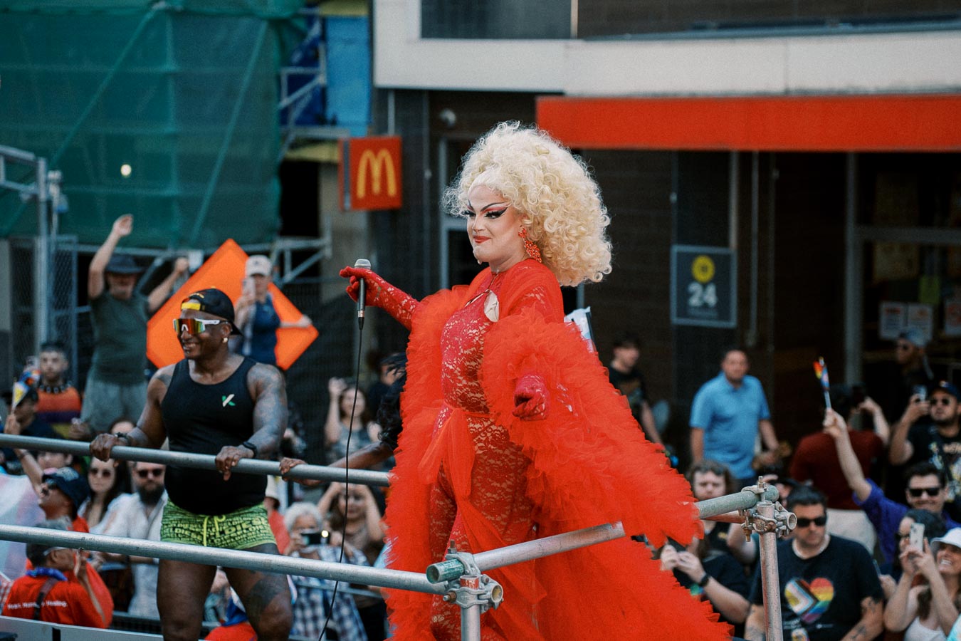 A drag performer in a vibrant red outfit and curly blonde hair participates in a lively parade, surrounded by a cheering crowd.