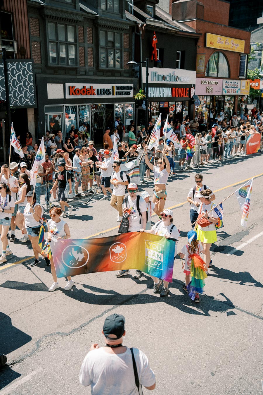 Participants march in a pride parade, holding a colorful banner and flags, with spectators watching from the sidewalk.