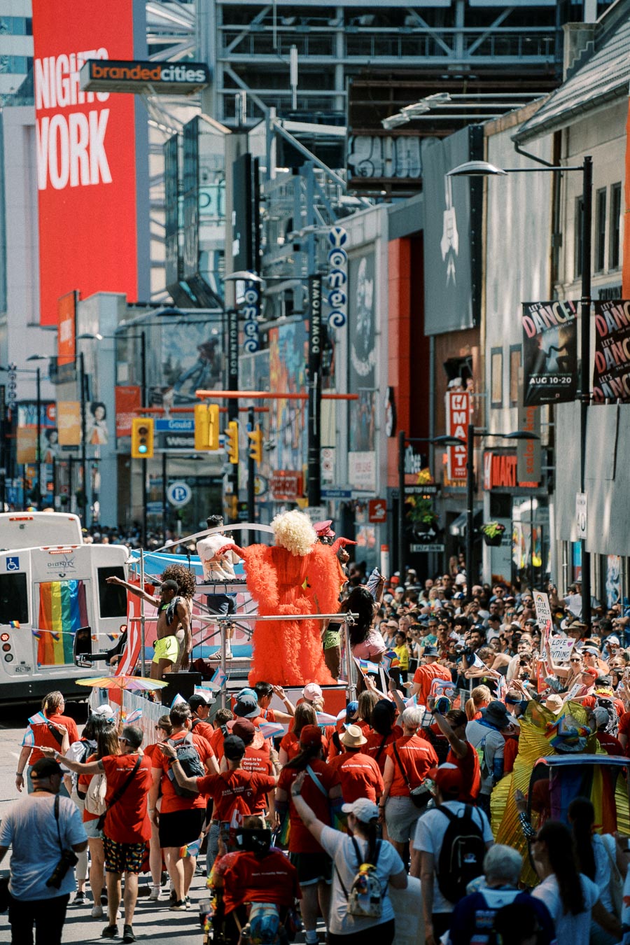 A lively street parade with colorful floats and performers draws a large crowd in an urban setting, showcasing vibrant costumes and festive energy.