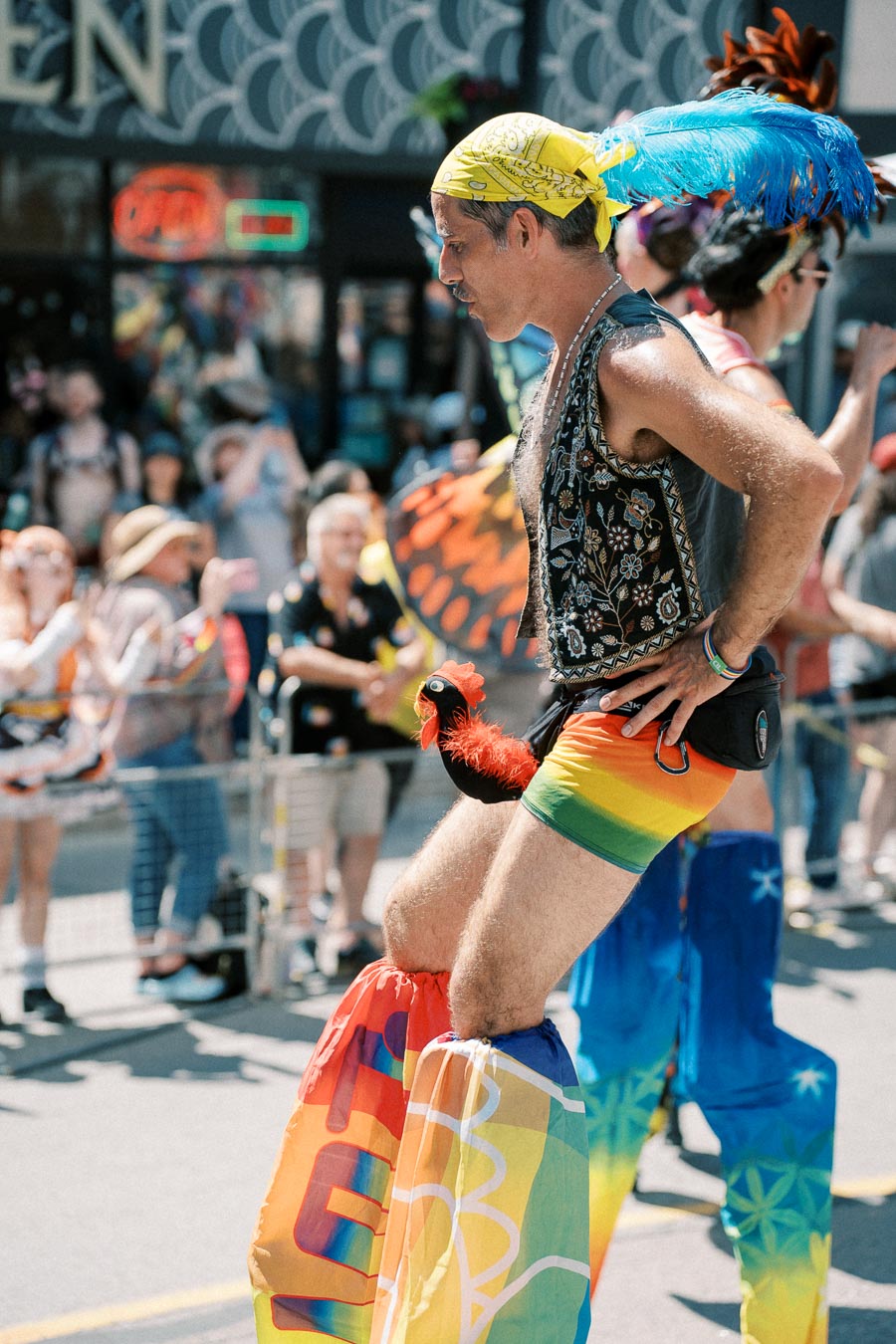 A person dressed in vibrant, colorful clothing and stilts participates in a lively parade, surrounded by a cheering crowd.