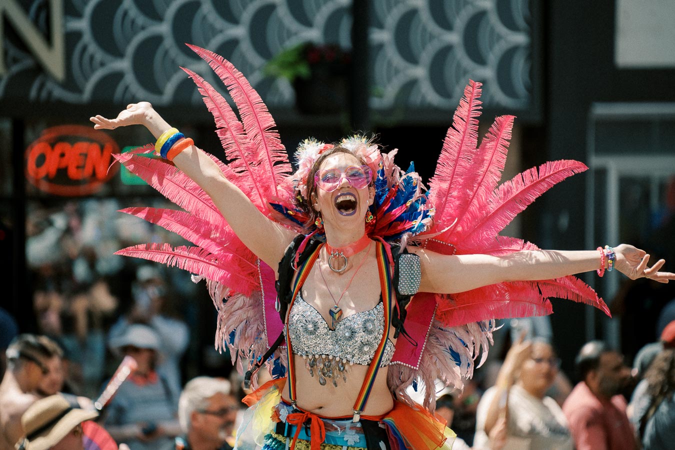 A joyful participant in a colorful costume with pink feathers and rainbow accessories at a lively parade.