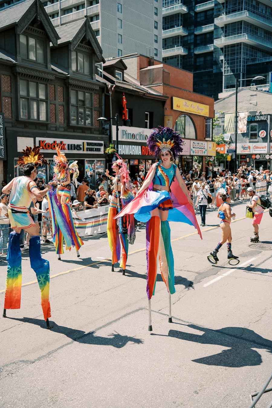 Participants in vibrant costumes and stilts take part in a lively parade on a city street, surrounded by spectators.