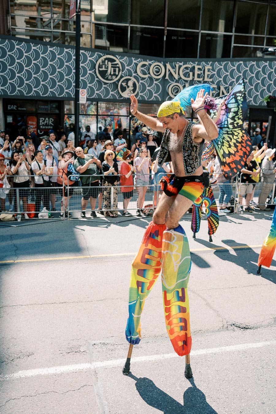 A vibrant parade participant on stilts, adorned with rainbow-colored attire and butterfly wings, entertains a cheering crowd.