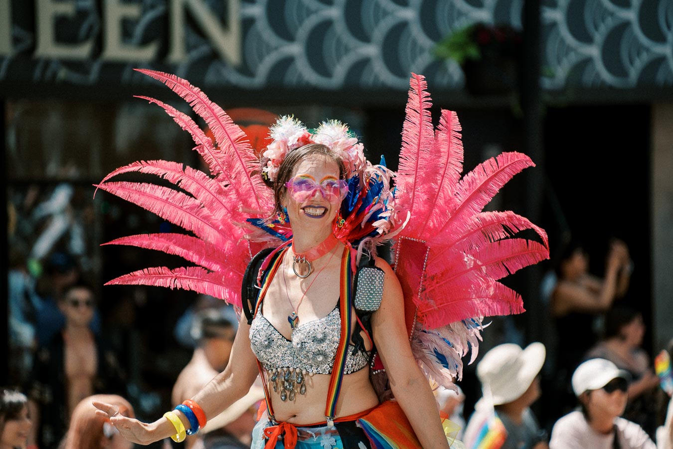 A person dressed in a vibrant costume with pink feathered wings and rainbow accessories participates in a parade, exuding joy and celebration.
