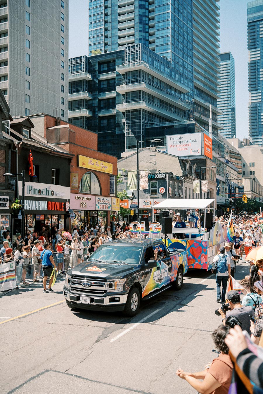 A vibrant parade takes place on a bustling city street, featuring a colorful float led by a Ford truck. Spectators line the sidewalks, enjoying the festive atmosphere amidst tall urban buildings.
