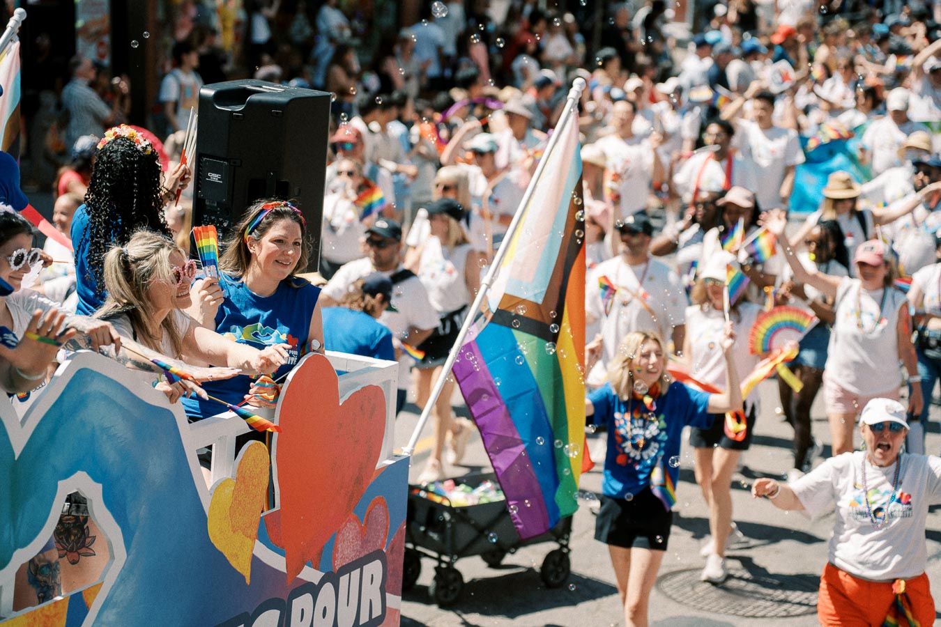 Participants celebrate at a pride parade, waving rainbow flags and wearing colorful attire, with a decorated float and a diverse crowd in the background.