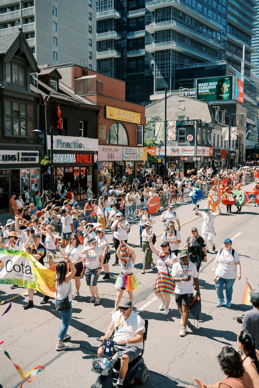 Participants in a vibrant parade march down a city street, celebrating with rainbow flags and colorful attire, while spectators line the sidewalks.