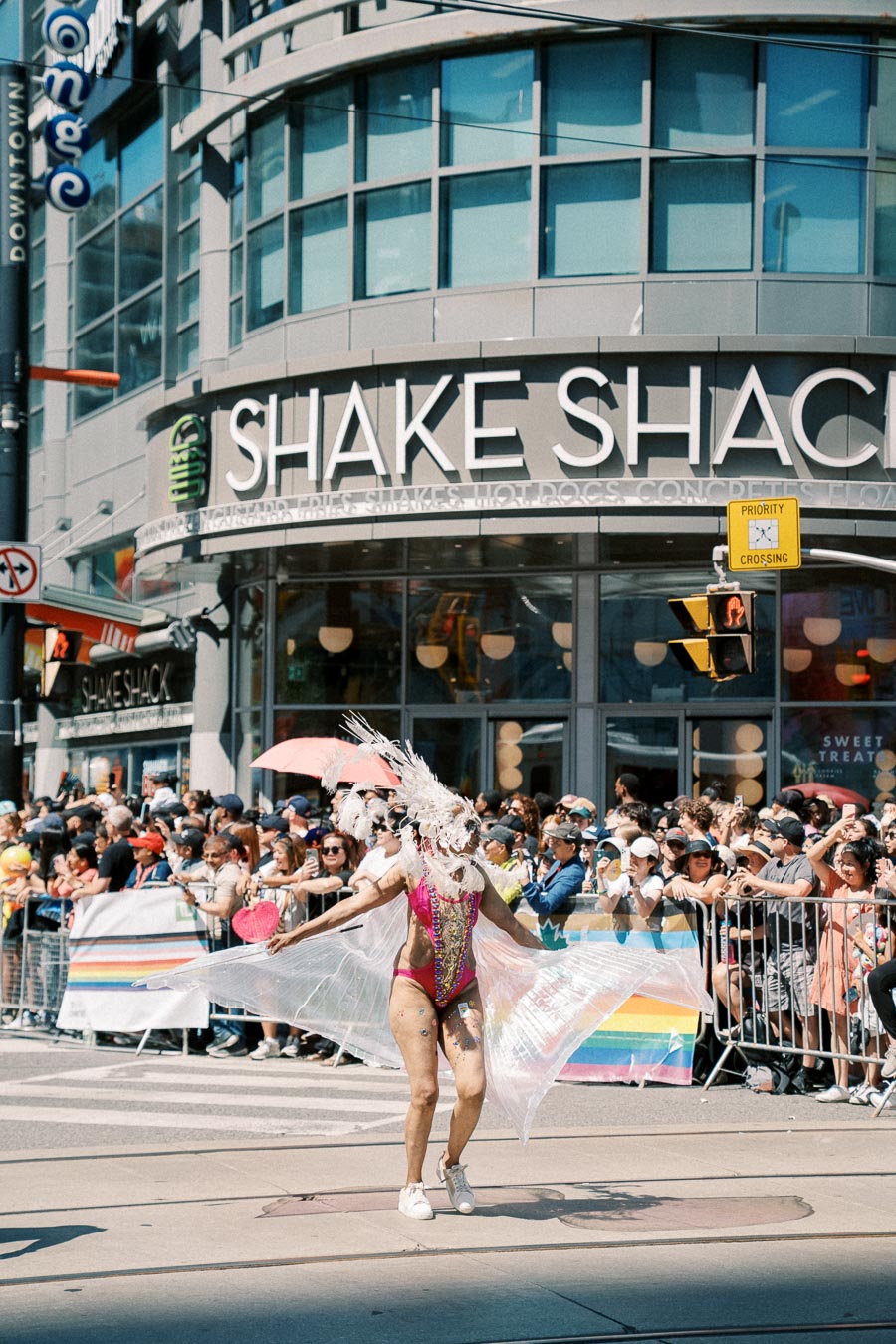 A vibrant parade participant in an elaborate costume walks past a Shake Shack, entertaining a large crowd gathered behind barriers.
