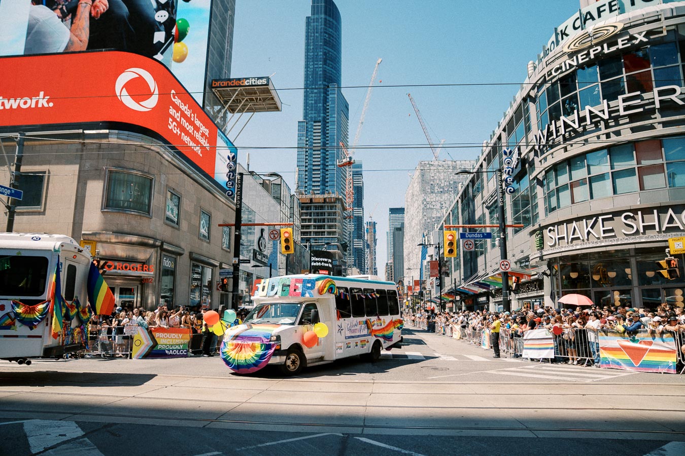 A lively pride parade in Toronto with a decorated bus and cheering crowds, set against the backdrop of the city's skyscrapers and digital billboards.