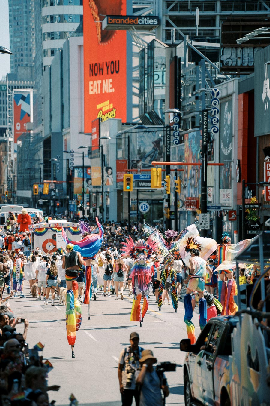 A lively parade featuring participants in colorful costumes and rainbow flags takes place on a bustling city street, with large advertisements and crowds lining the sidewalks.