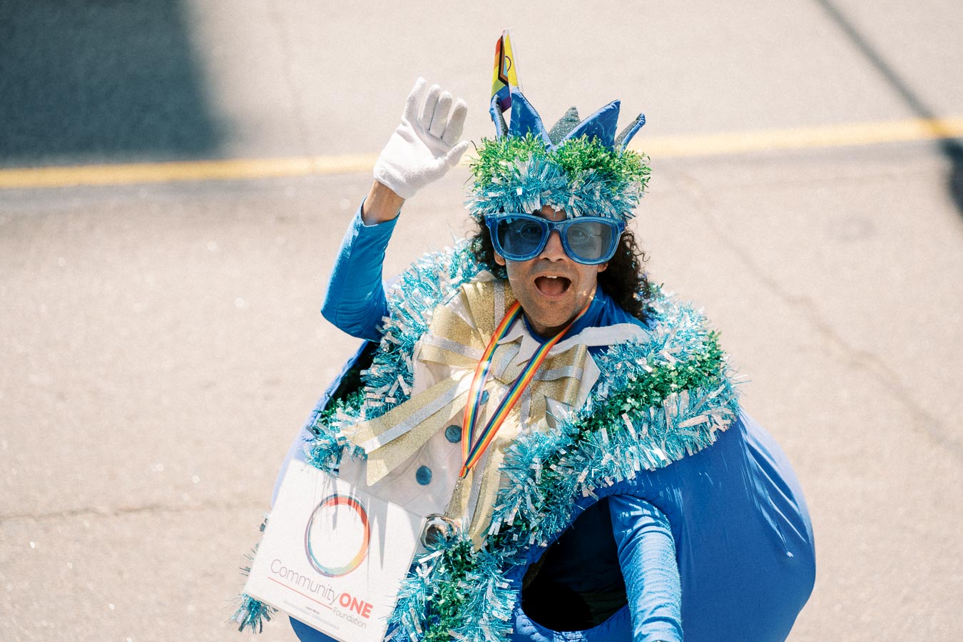A person dressed in a vibrant costume adorned with blue and green decorations waves enthusiastically during a parade. They are wearing large blue glasses and a white glove, and have a sign from Community One Foundation.