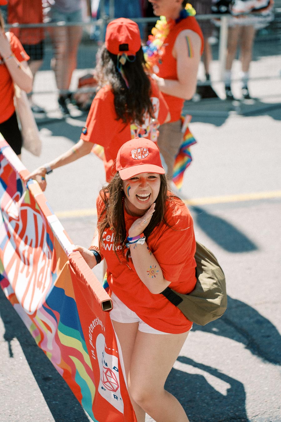 A participant in a parade wearing a red cap and shirt, holding a colorful banner, with rainbow face paint and a sun tattoo.