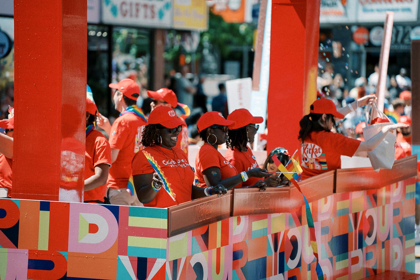 Participants in a pride parade wearing red shirts and hats, holding rainbow flags, and standing on a colorful float decorated with the word 'PRIDE'.