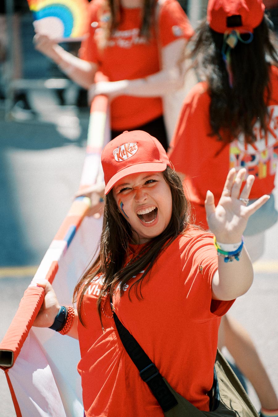A participant in a parade enthusiastically waves while holding a rainbow flag, wearing a red KitKat cap and shirt.
