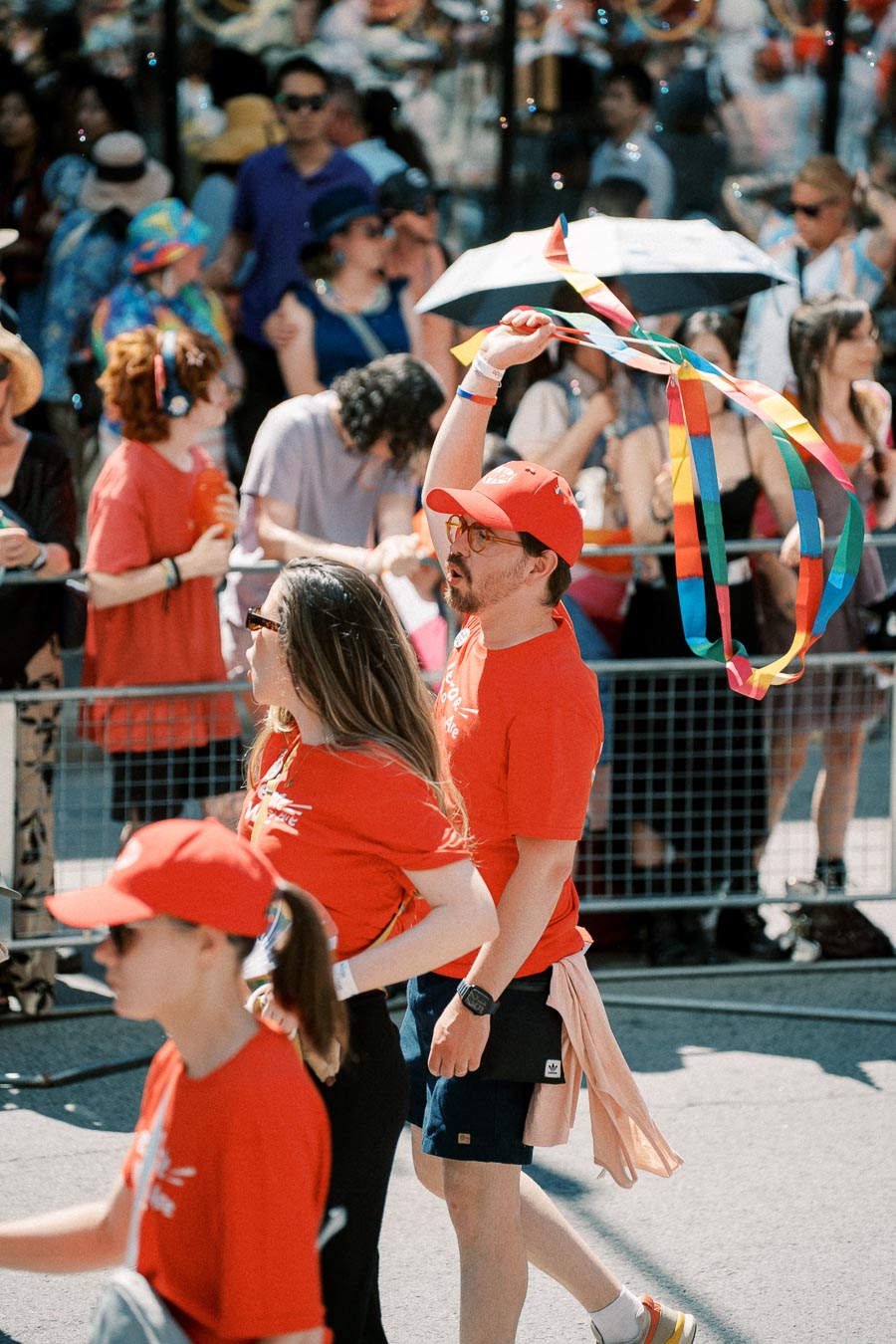 Participants in a parade wearing red shirts and hats, with one person waving rainbow ribbons, surrounded by a crowd.