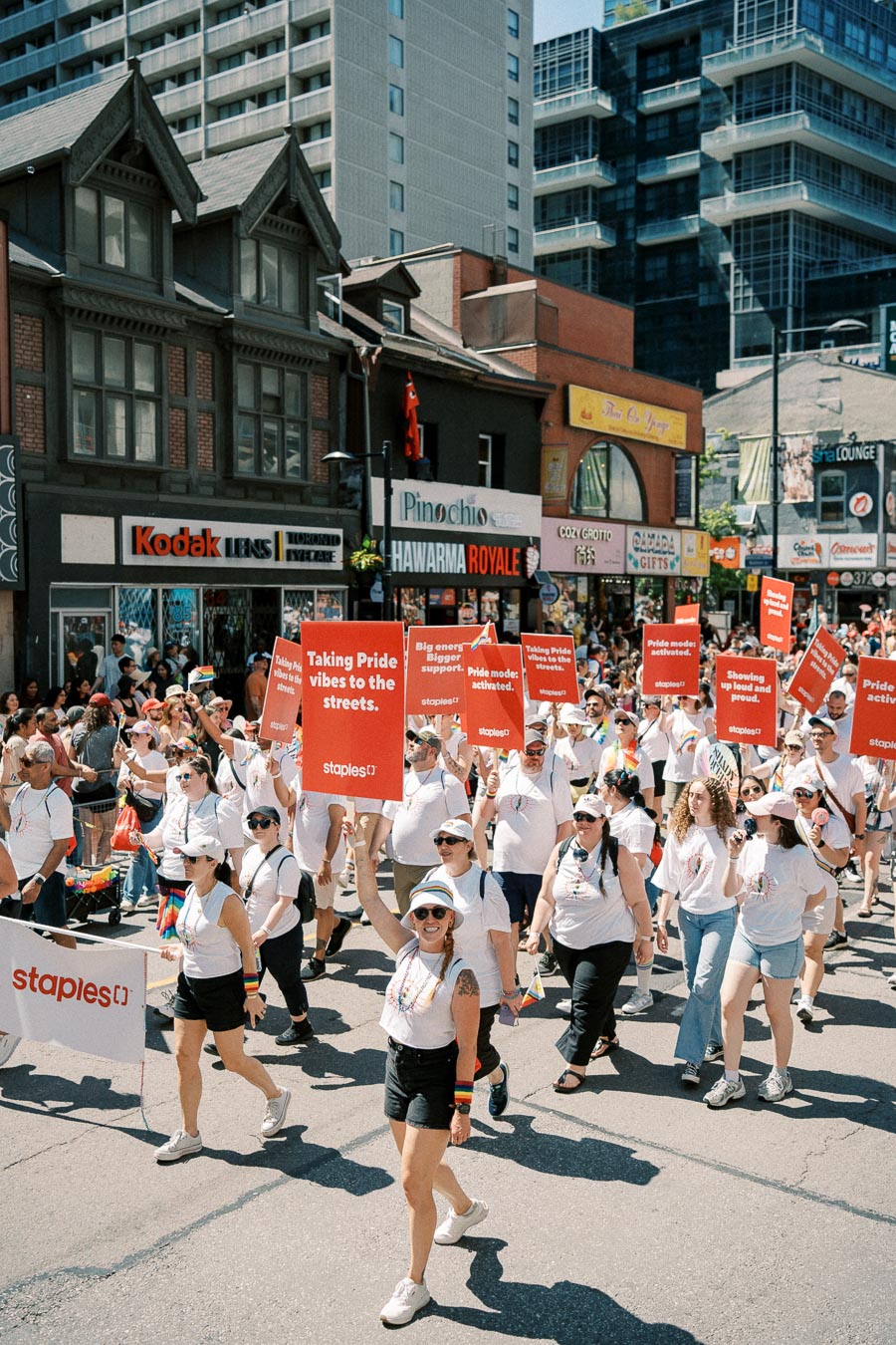 Participants in a Pride parade march down a city street, holding signs with supportive messages. The parade features people in white shirts, celebrating diversity and inclusion.