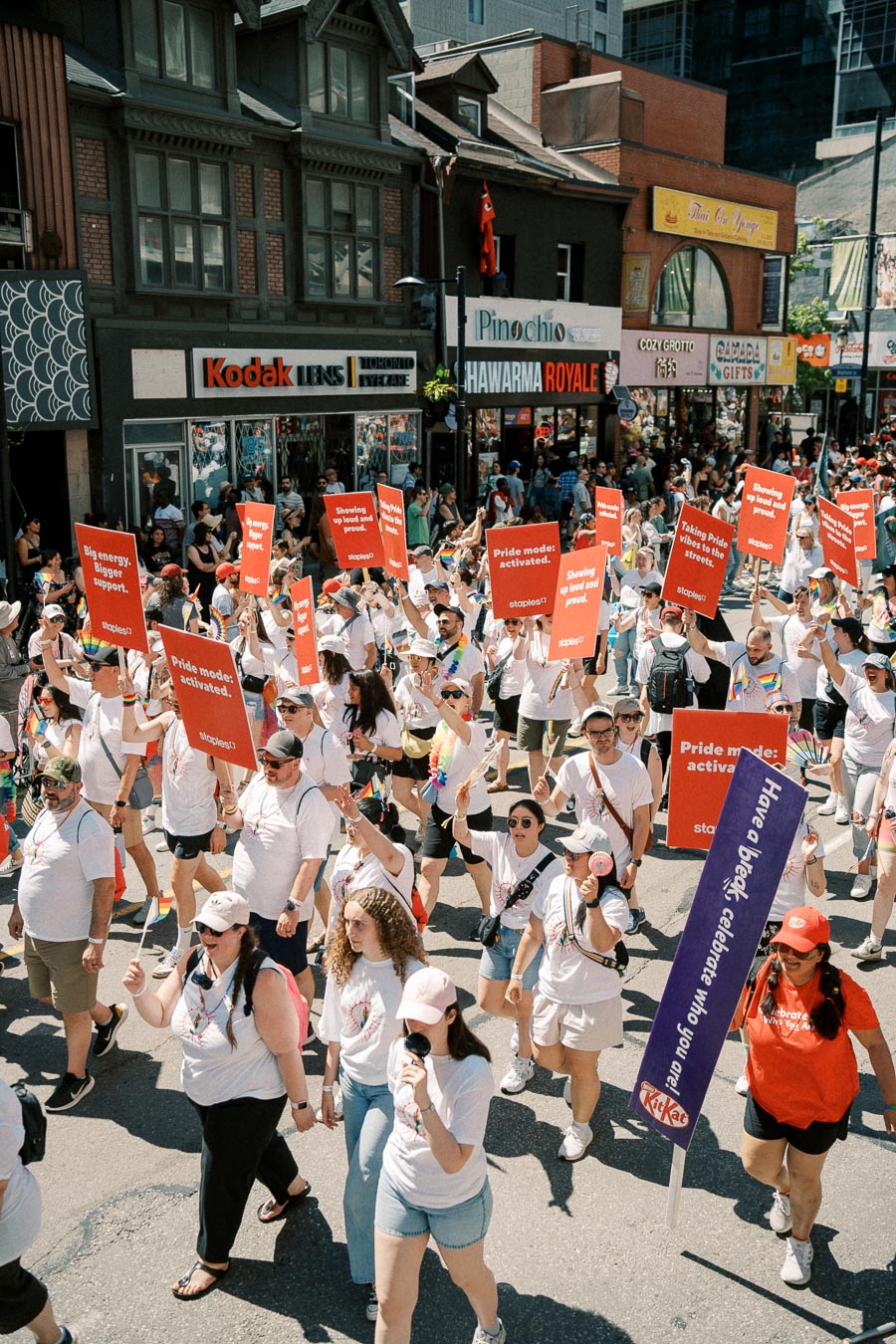 Participants in a pride parade march down a city street, holding signs with messages of support and pride. The street is lined with various shops and spectators.