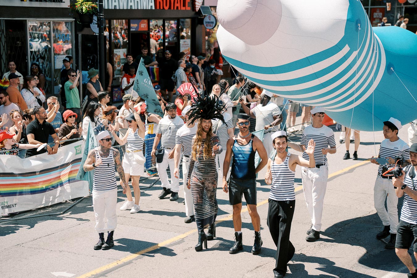 Participants in sailor outfits march in a vibrant parade, accompanied by a large inflatable object, while spectators cheer from the sidelines.