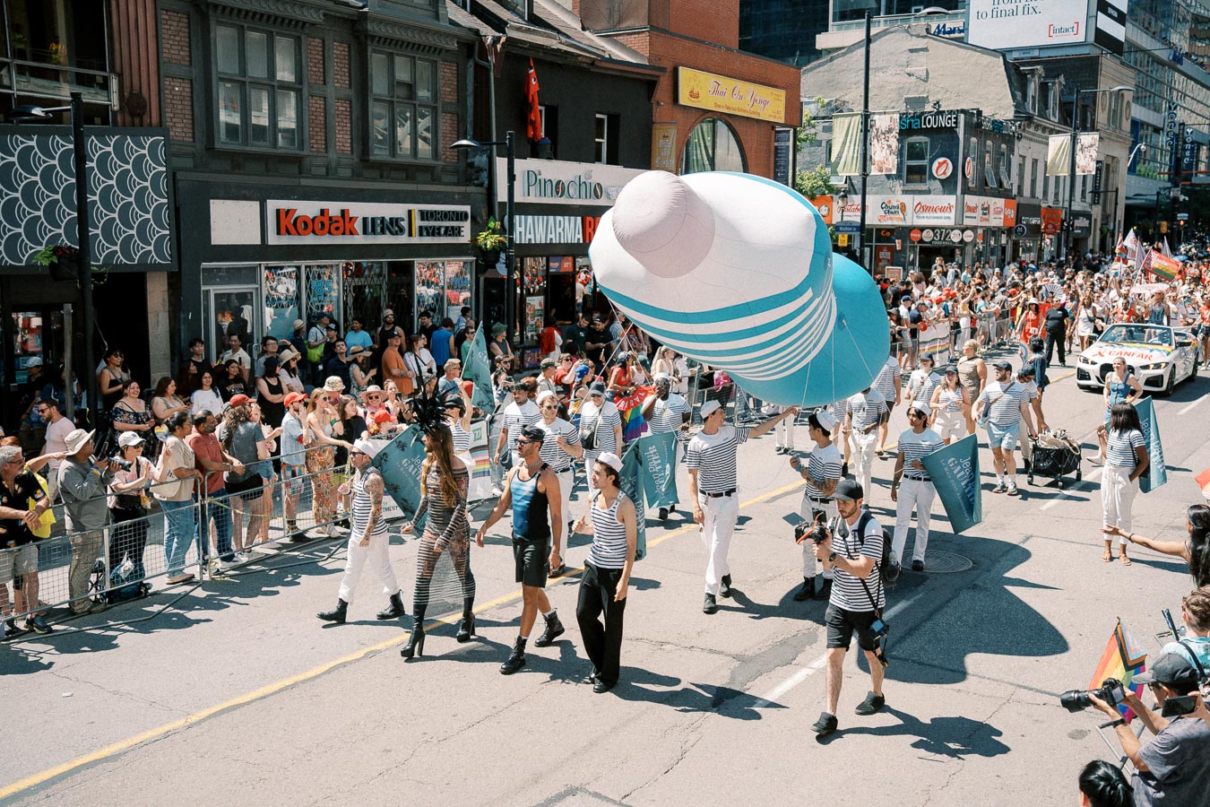 Participants in sailor outfits march in a vibrant street parade, carrying a large inflatable object, while spectators watch from the sidelines.