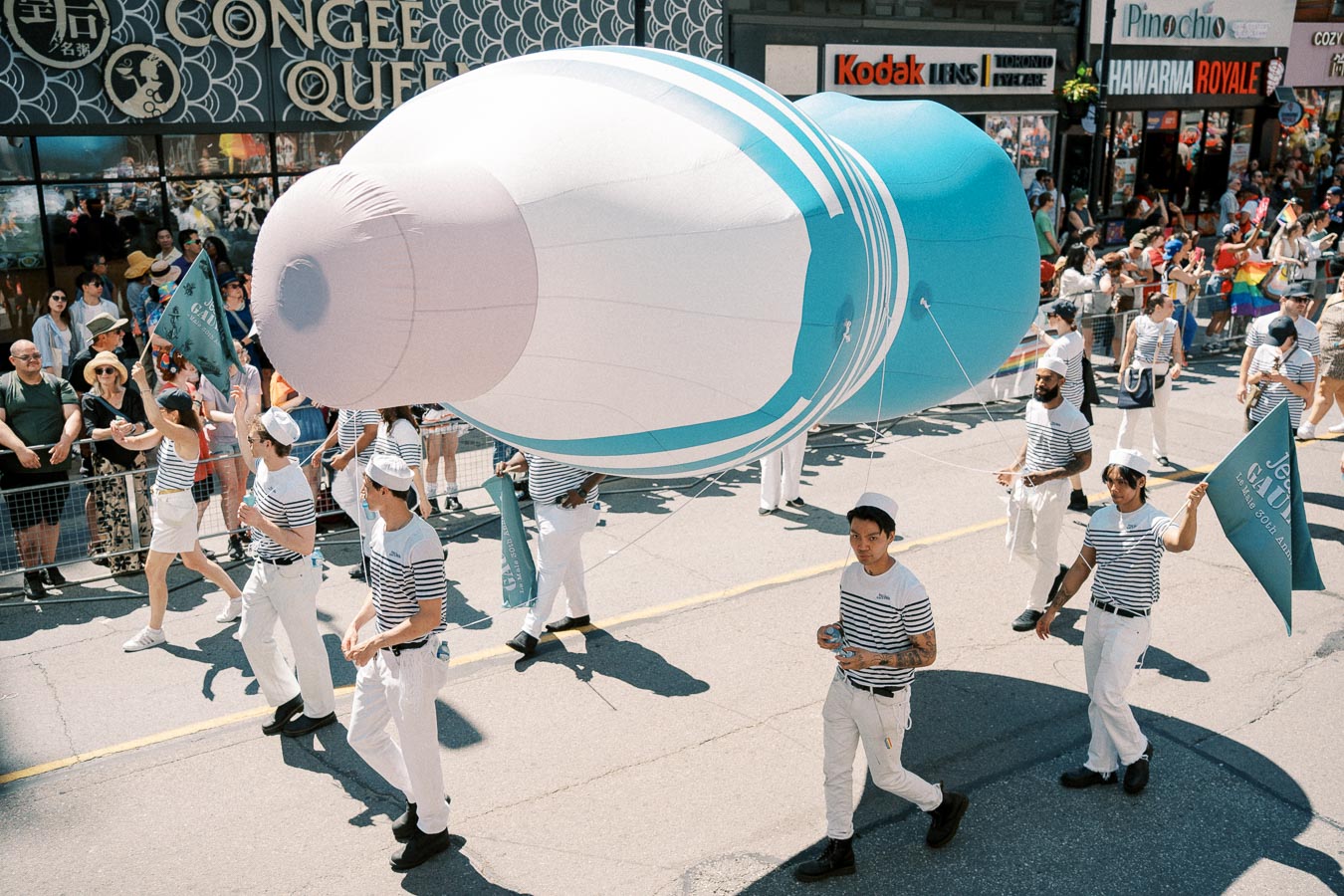 Participants in sailor outfits carry a large inflatable object resembling a bottle during a parade, with spectators watching from the sidelines.
